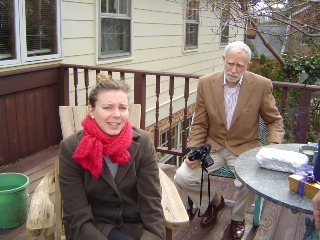 Valerie (visting from Switzerland) all bundled up wearing a bright red scarf while seated on a wooden adirondeck chair, while 	Uncle Paul looks on with camera in hand.