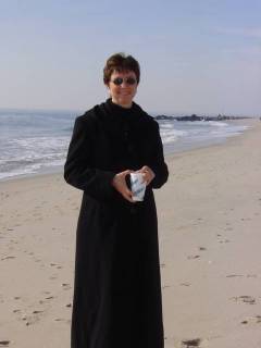 Ann wearing a long black coat, standing in front of the ocean holding a styrofoam cup of hot tea.